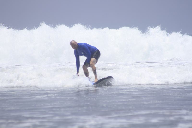 Beginner surfer in a blue rash vest crouching low as he rides a soft-top board through the whitewater