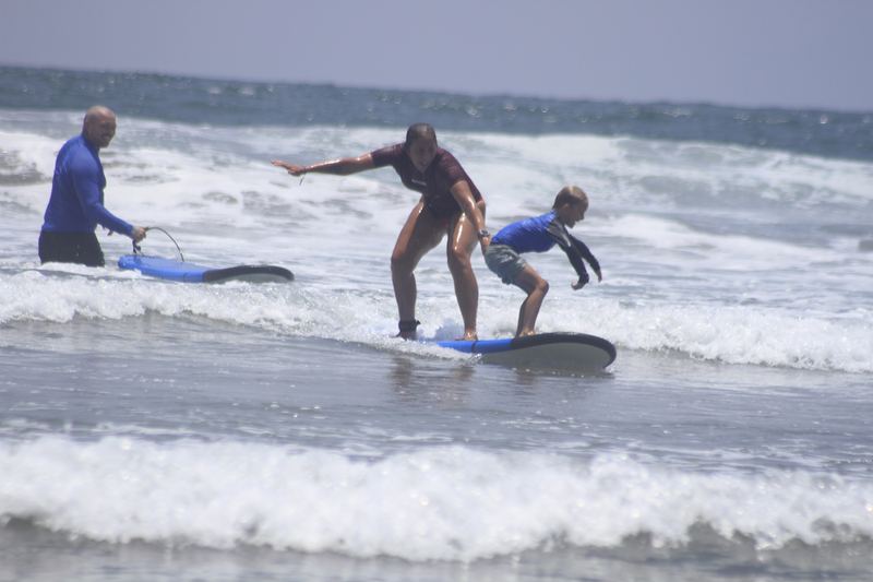 Two learners sharing a soft-top board in the whitewater while another surfer in a blue rash vest stands nearby with his board