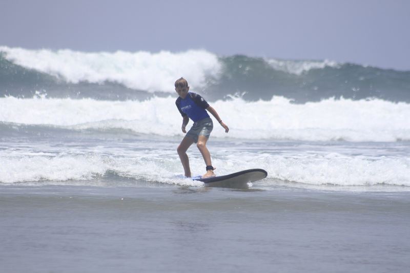 Young beginner surfer in a blue rash vest standing up on a soft-top board in the whitewater